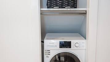 A compact laundry room with a front-loading washing machine and a shelf above it containing cleaning supplies.