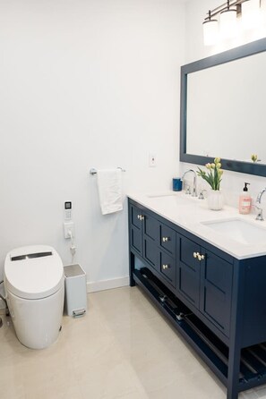 Ein modernes Badezimmer mit Toilette, Mülleimer, Waschbecken mit Spiegel und einer kleinen Pflanze auf der Ablagefläche.
