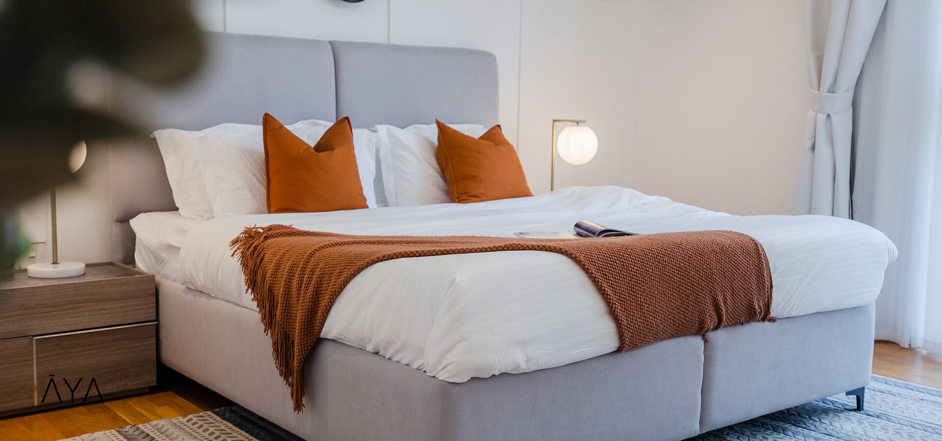 Ein modernes Schlafzimmer mit einem grauen Bett, weißer Bettwäsche und zwei orangefarbenen Kissen. Ein Nachttisch mit Lampe und einer kleinen Schublade ist ebenfalls vorhanden.