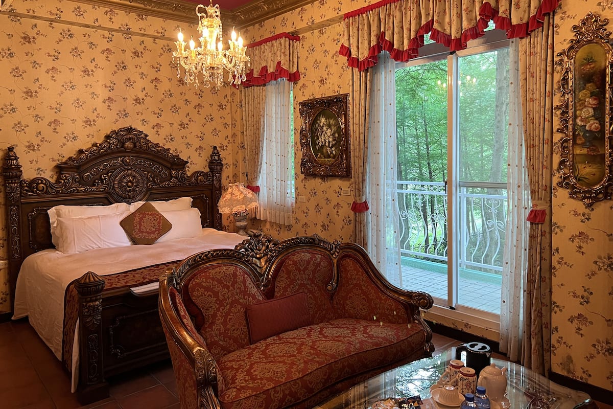 A bedroom with a large ornate bed, a chaise lounge, a chandelier, and a view of a balcony.