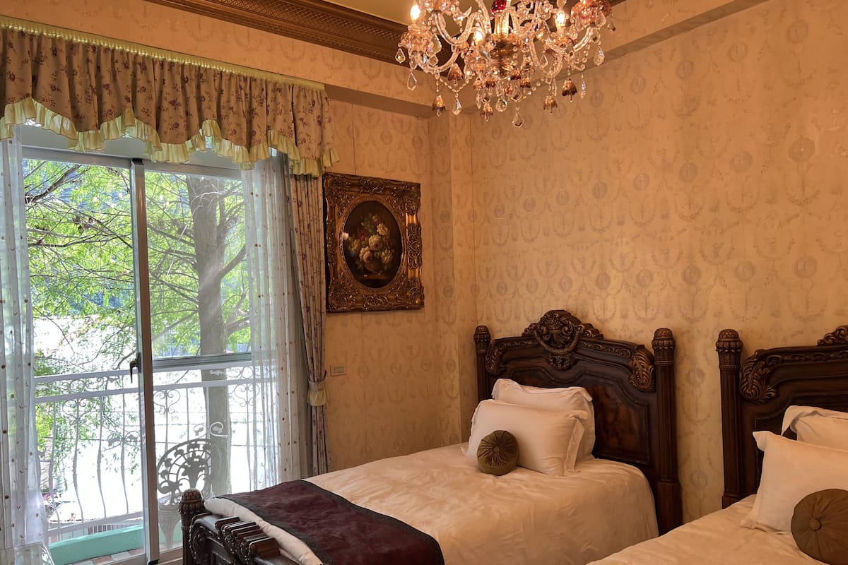 A bedroom with two beds, a chandelier, a window with curtains, and a framed picture on the wall.