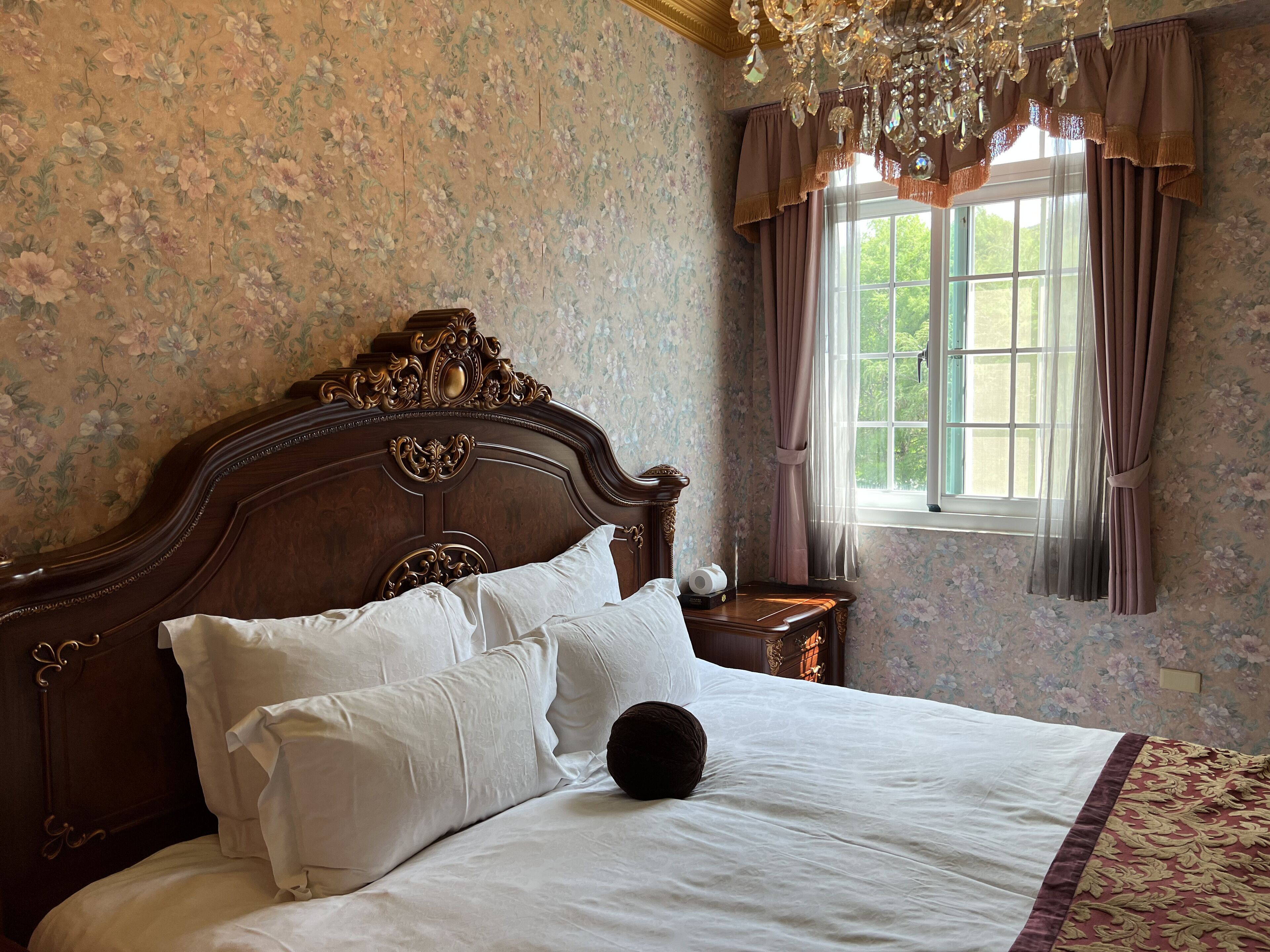 A bedroom with a wooden bed, floral wallpaper, a chandelier, and a window with curtains.