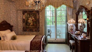 Une chambre classique avec un grand lit, un lustre, un tableau et une vue sur la verdure par la fenêtre.