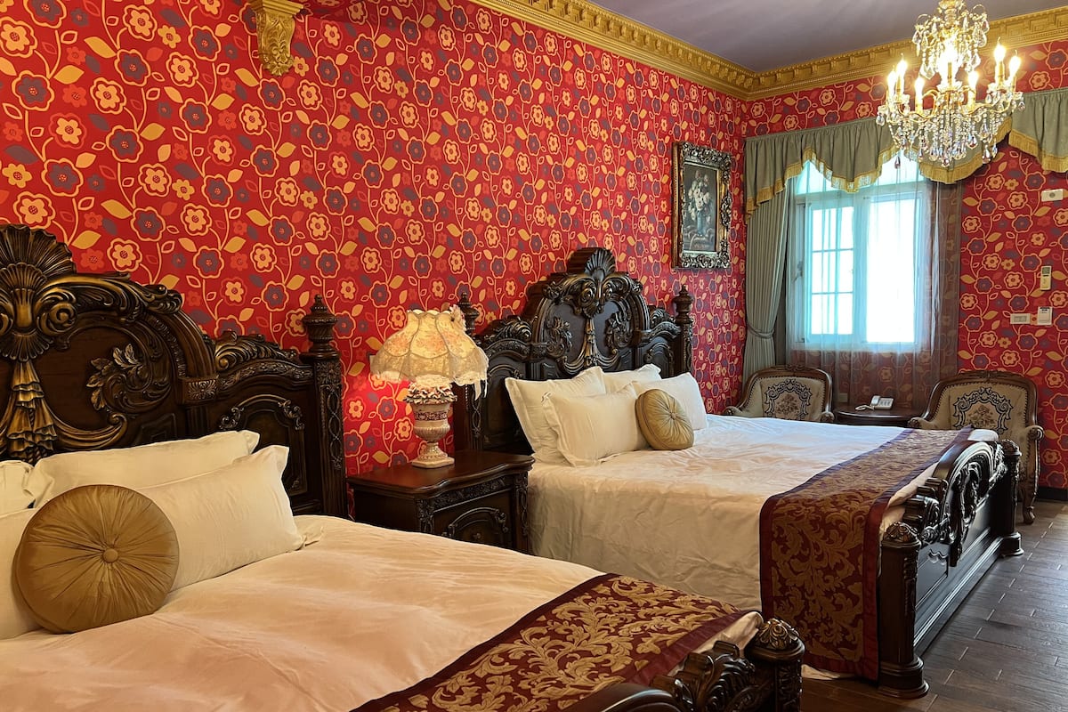 A room with two ornate beds, a chandelier, and floral wallpaper.