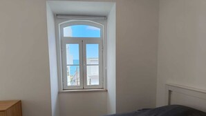 A bedroom with a single bed, a window with a view of buildings and the sea, and a wooden dresser. - Beach Haven (Albufeira)