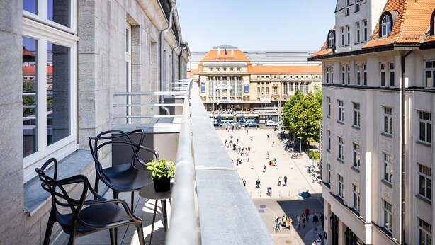 Un balcone con due sedie e un tavolino, affacciato su una piazza con edifici e un trammo sullo sfondo.