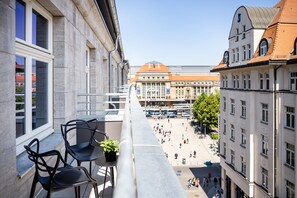 A balcony with two chairs and a small table, overlooking a public square with buildings and a tram in the background. - AMANO Home Leipzig (Leipzig)
