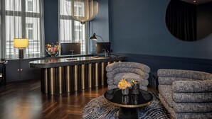 Un soggiorno moderno con una parete blu scuro, un tavolino rotondo, due poltrone a fantasia, un'ampia finestra con tende leggere, un lampadario e un televisore su un mobile.