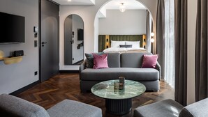 Camera d'albergo moderna con divano, tavolino rotondo in vetro, televisore e letto con testiera.