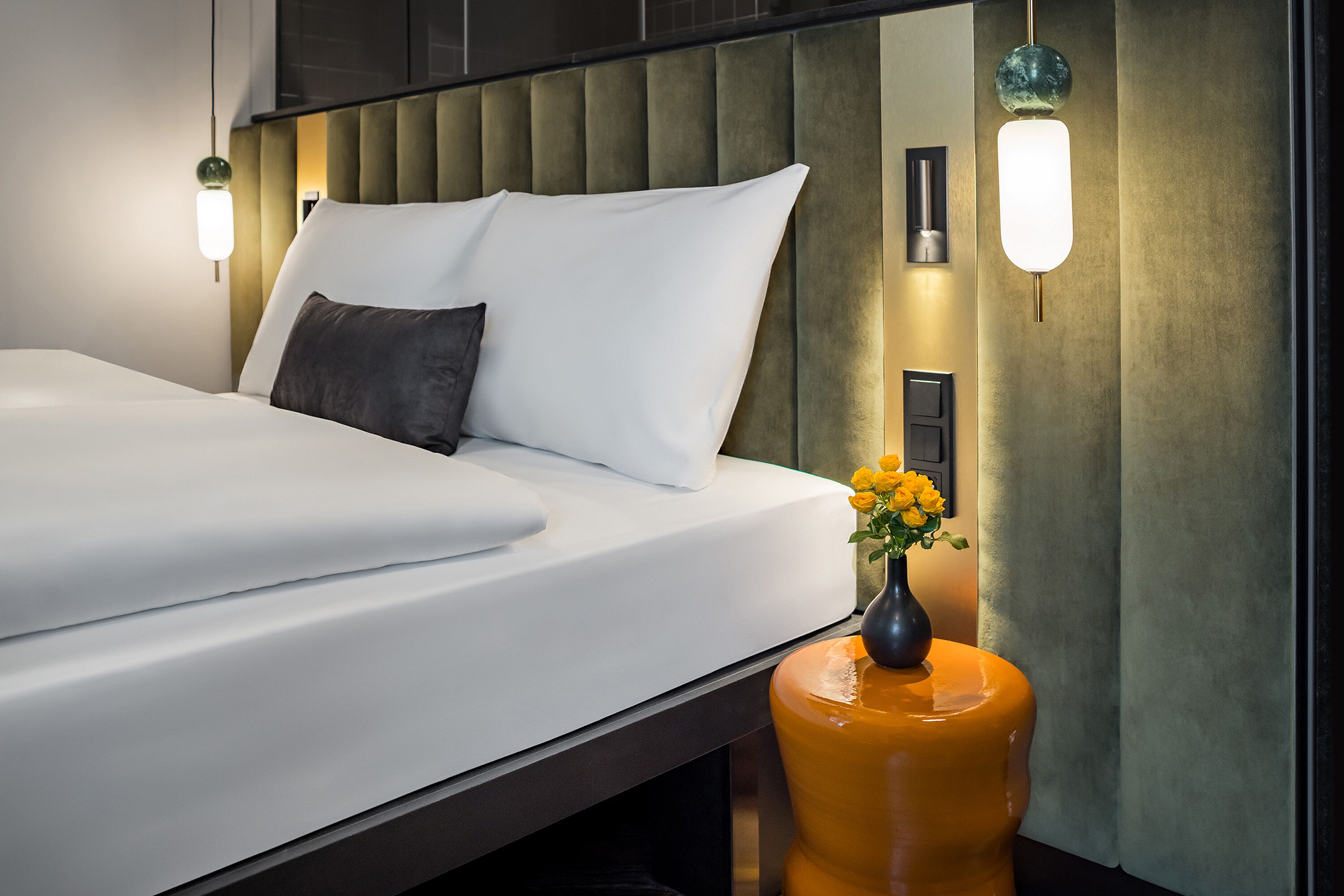 A modern hotel room with a neatly made bed, a vase of yellow flowers, and ambient lighting.