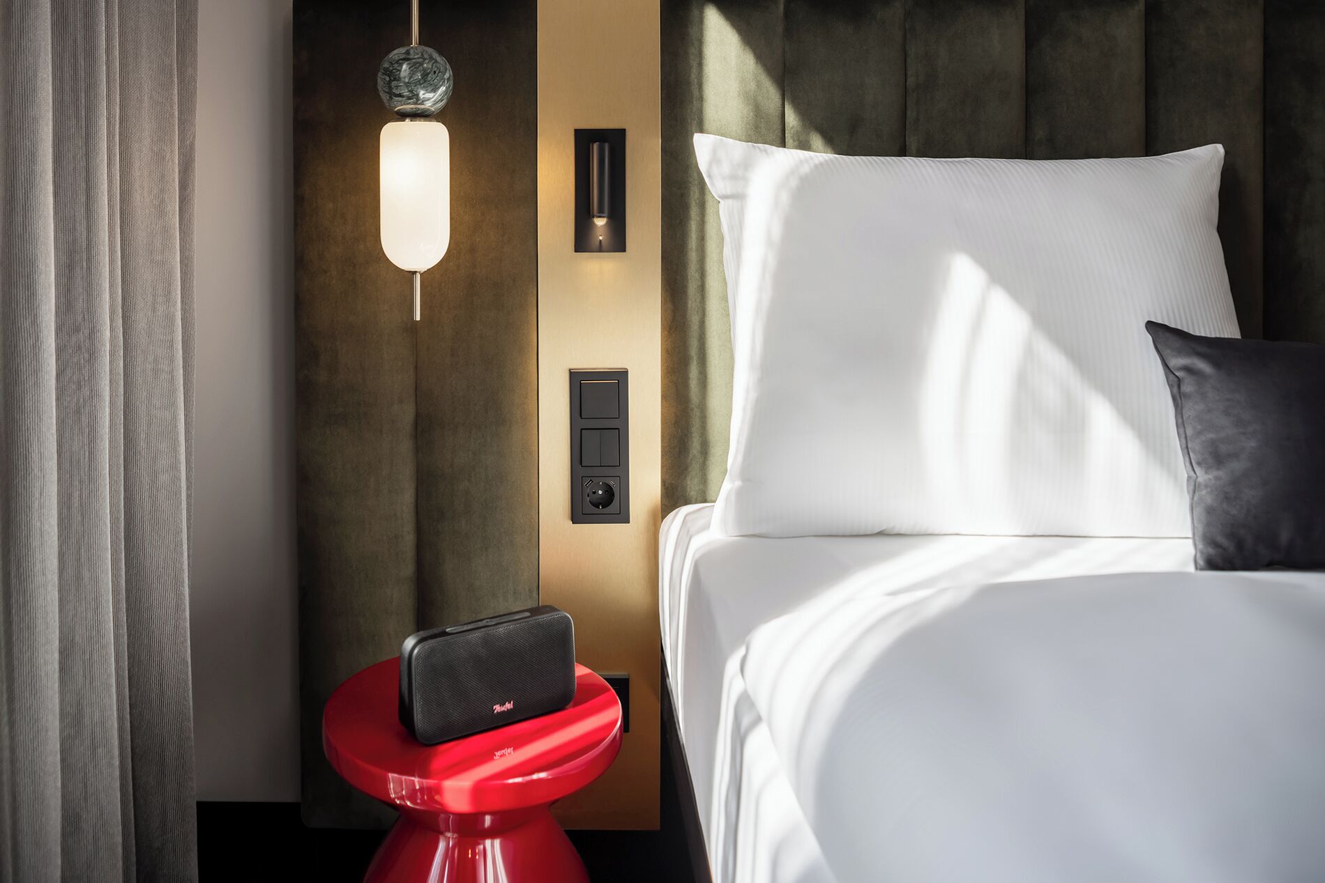 A hotel room with a bed, a red side table, a wall-mounted control panel, and a hanging light fixture.