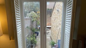 Une double porte-fenêtre donne sur une cour arrière agrémentée d’un mur en briques, de plantes en pot et d’une clôture en bois.