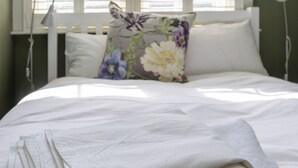 Un lit bien fait, avec du linge de lit blanc, un coussin à motifs floraux et, en arrière-plan, une fenêtre avec des stores.