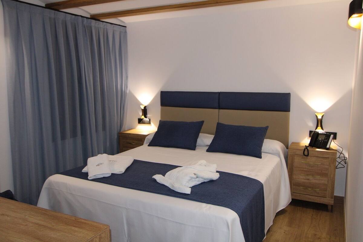 A hotel room with a bed, two bedside tables, a wooden nightstand, a desk, and a chair.