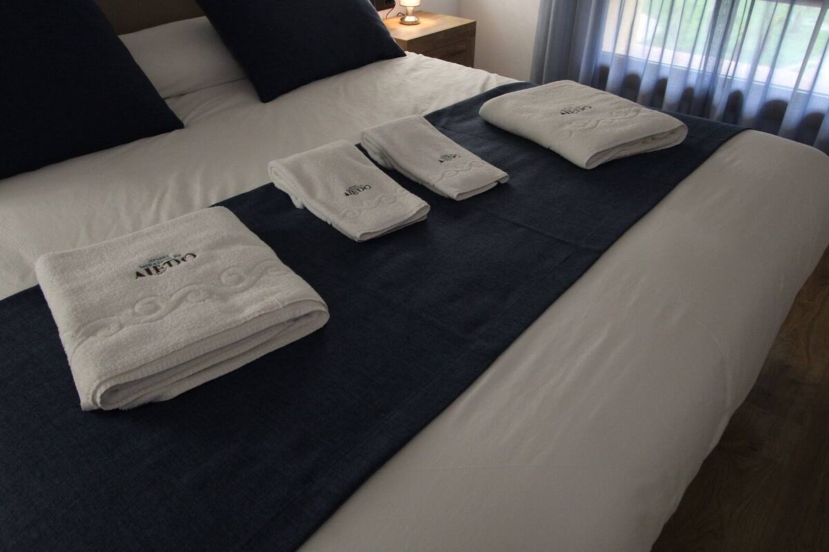 A neatly made bed with white linens and a dark blue bedspread, accompanied by folded white towels with a logo.