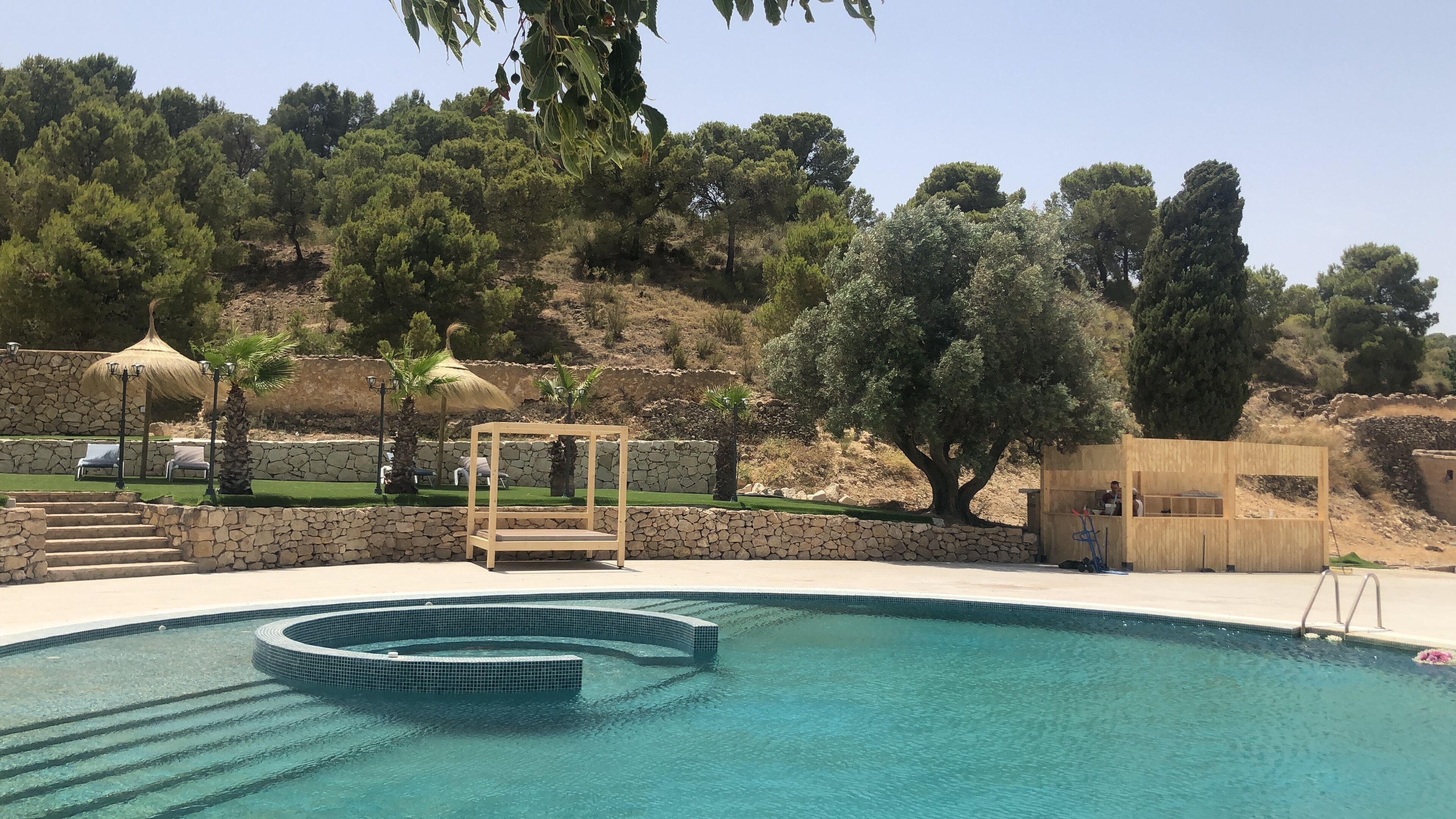 Una piscina con un muro de piedra, un pabellón de madera y una pequeña construcción de madera.