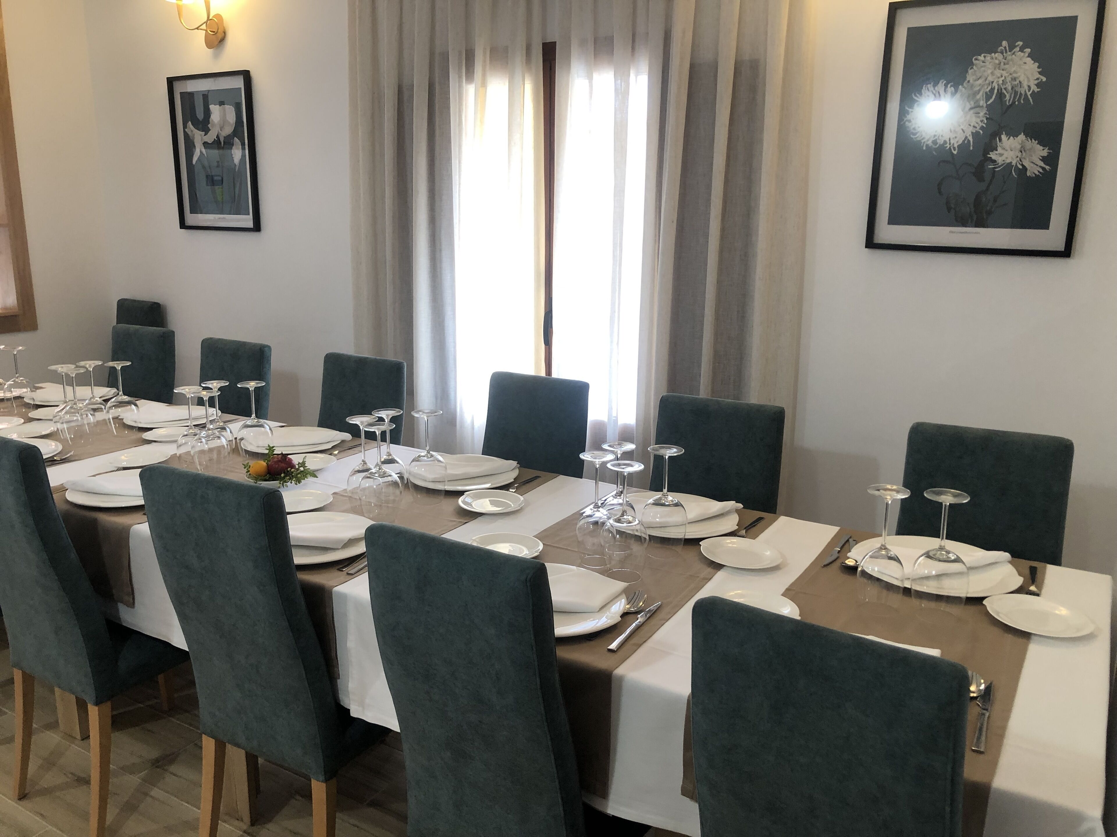 Une salle à manger avec une longue table dressée pour un repas, entourée de chaises, et des œuvres d’art encadrées accrochées aux murs.