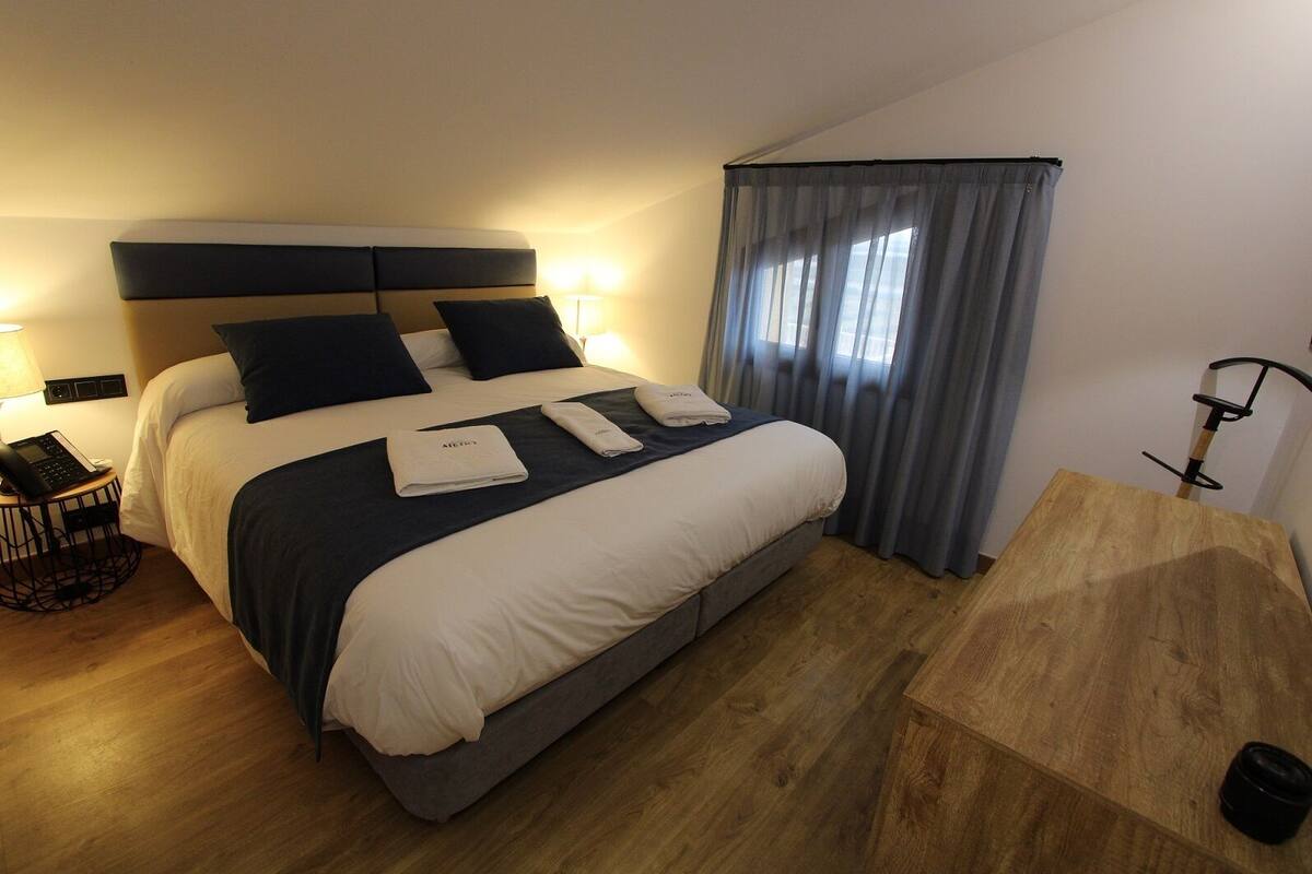 A neatly made bed with a grey headboard, a bedside table with a lamp, and a wooden desk with a chair.