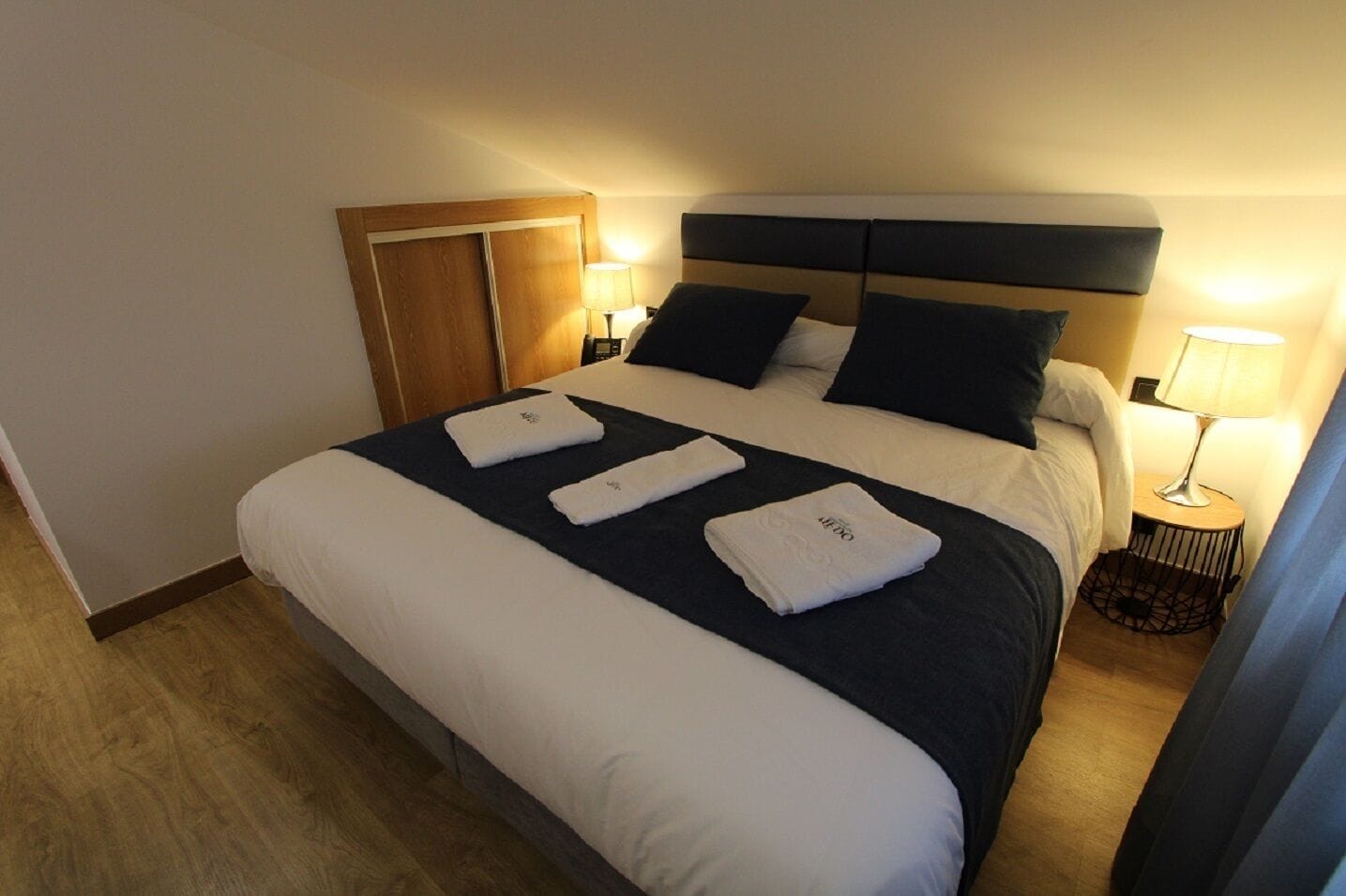 Una cama bien hecha con ropa de cama blanca y azul oscuro, un armario de madera y dos lámparas de noche.