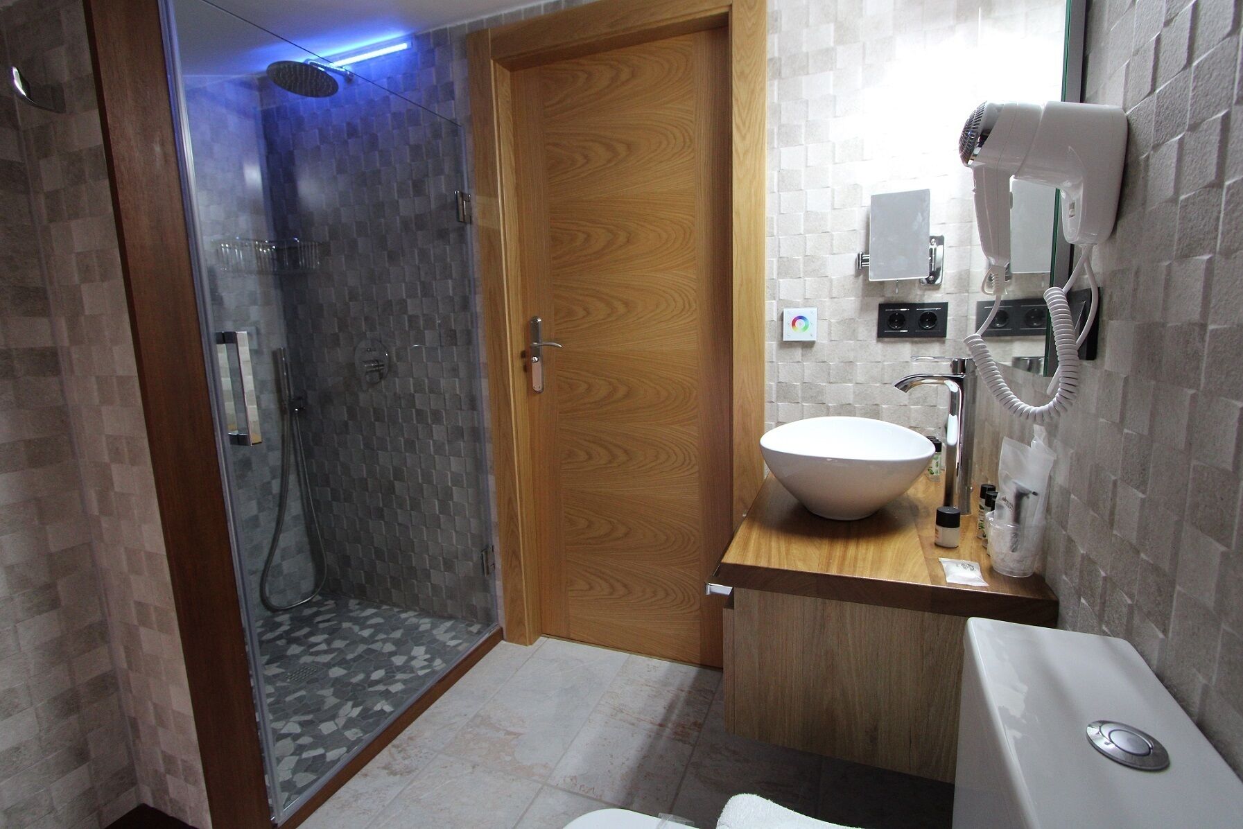 Un baño moderno con ducha acristalada, puerta de madera, lavamanos blanco y secador de pelo montado en la pared.