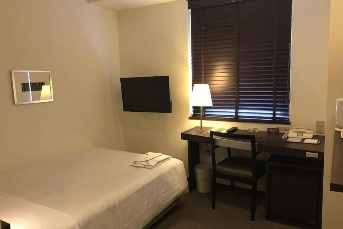 Une chambre d’hôtel équipée d’un lit, d’un bureau, d’une chaise, d’une télévision, d’une lampe et de stores aux fenêtres.