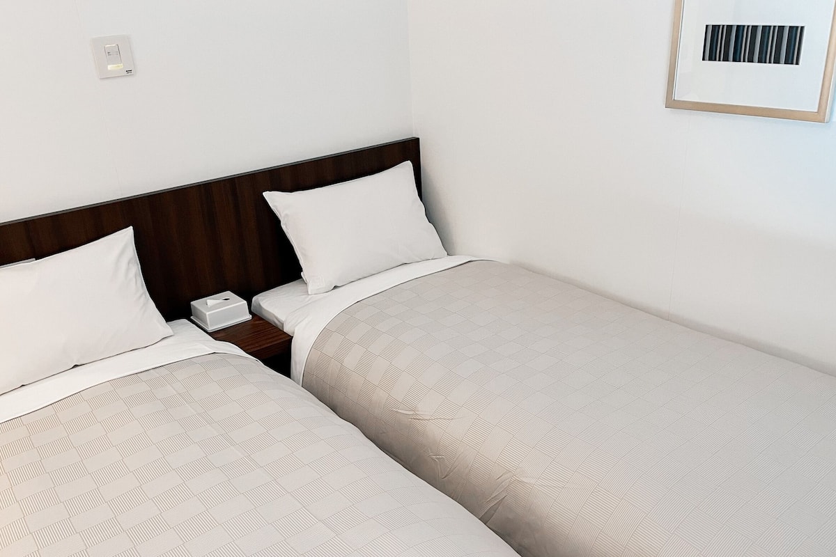 Une chambre d’hôtel avec deux lits simples, chacun doté d’une tête de lit en bois, de draps blancs et d’une table de chevet avec un téléphone et une boîte de mouchoirs.