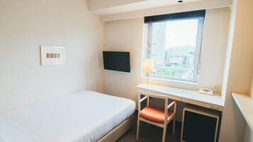 Quarto de hotel com uma cama de solteiro, uma escrivaninha com cadeira, televisão na parede e janela com vista para edifícios.
