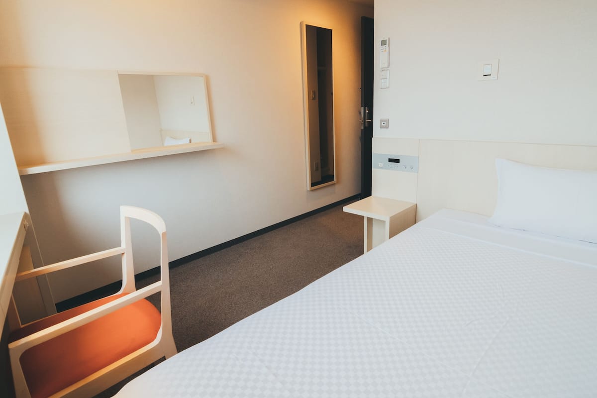 Une chambre d’hôtel avec un lit, une chaise dotée d’un coussin orange, une petite table, un miroir et un panneau de commande fixé au mur.