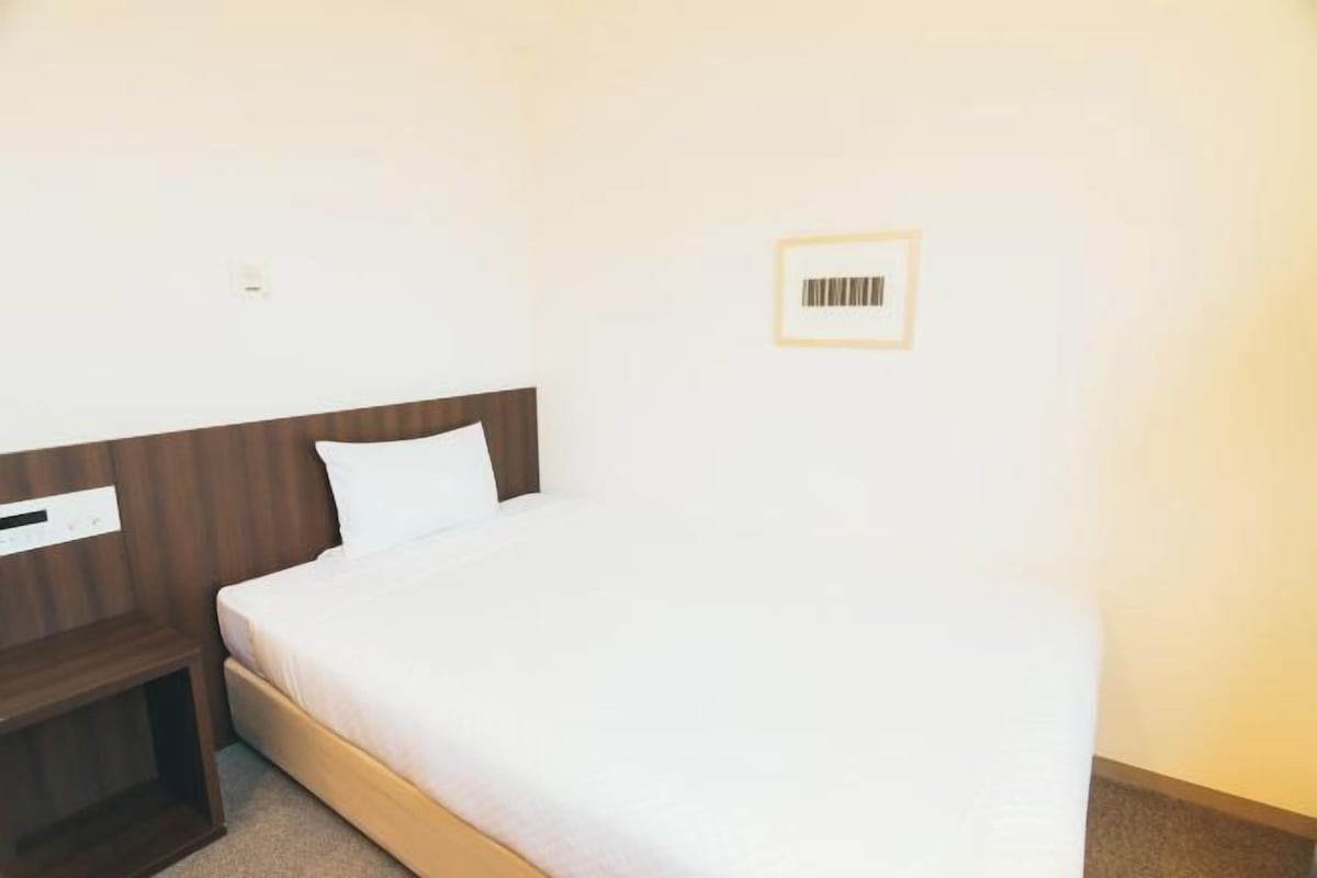 Una cama individual con cabecera de madera en una habitación de paredes blancas y una mesita de noche pequeña y sencilla.