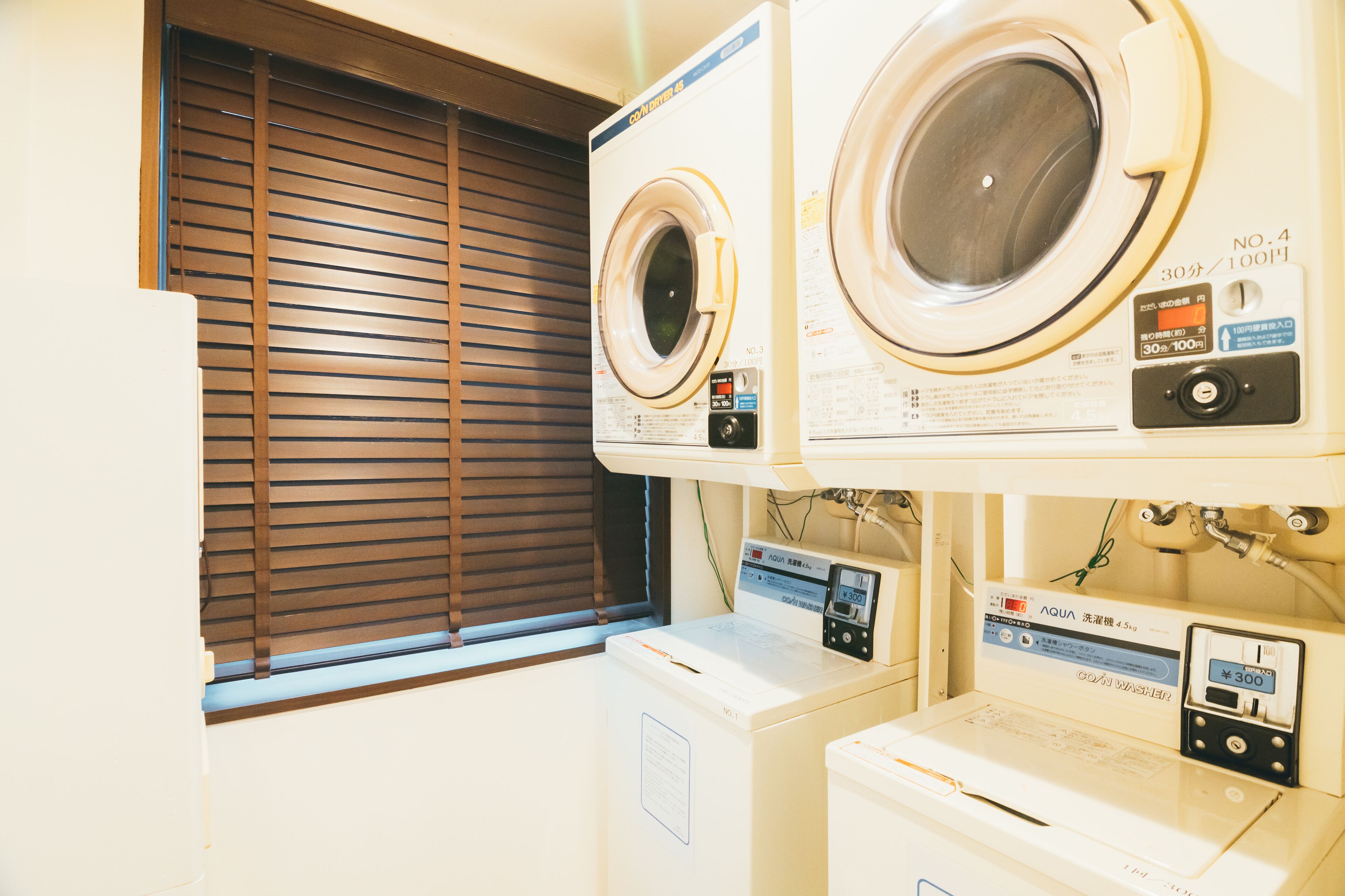 Un espace buanderie équipé de deux machines à laver et d’un sèche-linge, une fenêtre avec des stores et un panneau de commande sur les appareils.