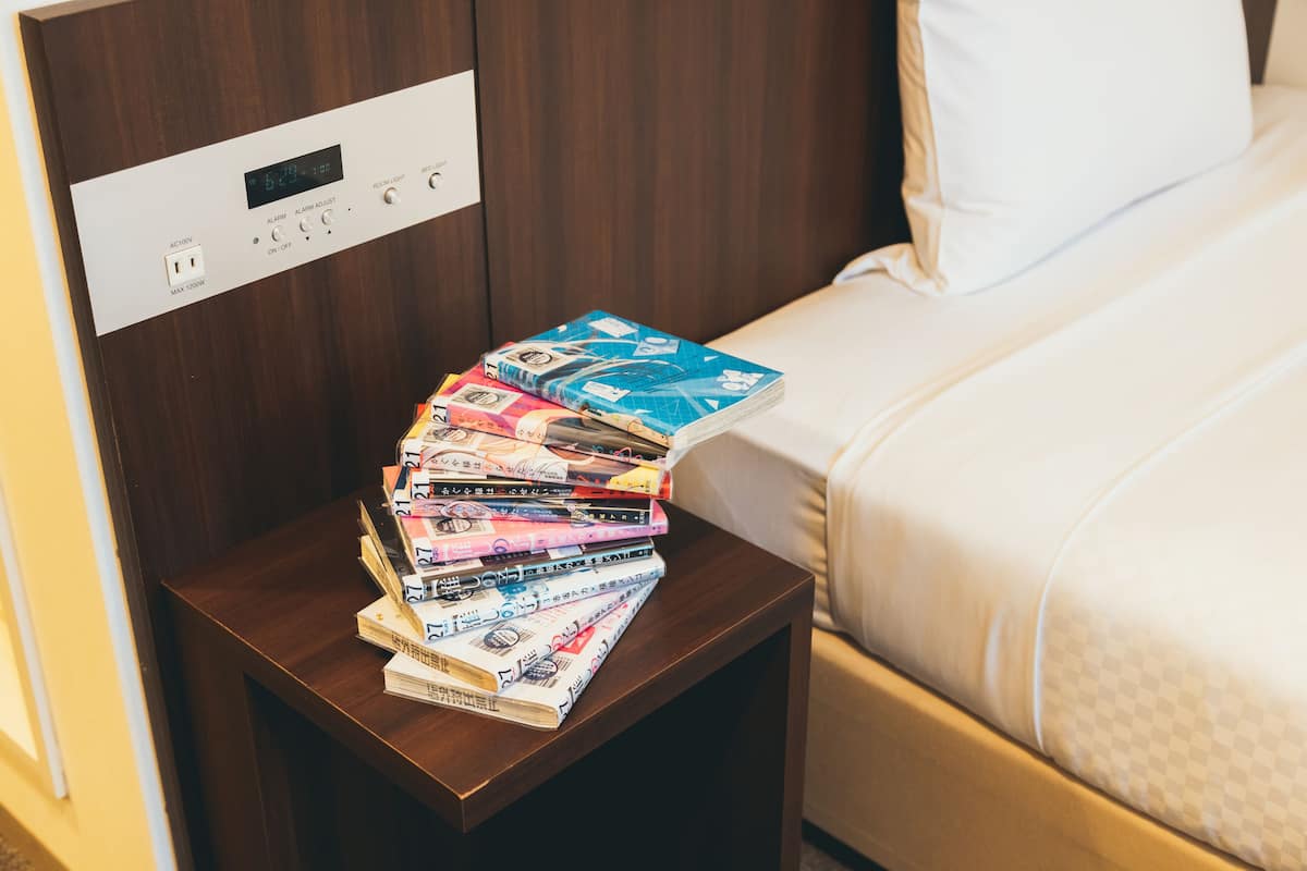 Une pile de billets de banque sur une table de chevet en bois, à côté d’un lit avec un oreiller blanc.