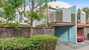 Un bâtiment moderne doté d’une grande fenêtre, d’un balcon orné de plantes en pot et d’une voiture rouge garée dans l’allée.
