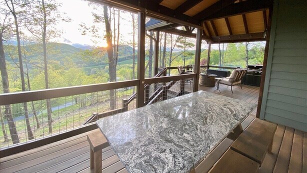 Una terraza techada al aire libre con una mesa de mármol, barandas de madera y vistas a un paisaje boscoso.