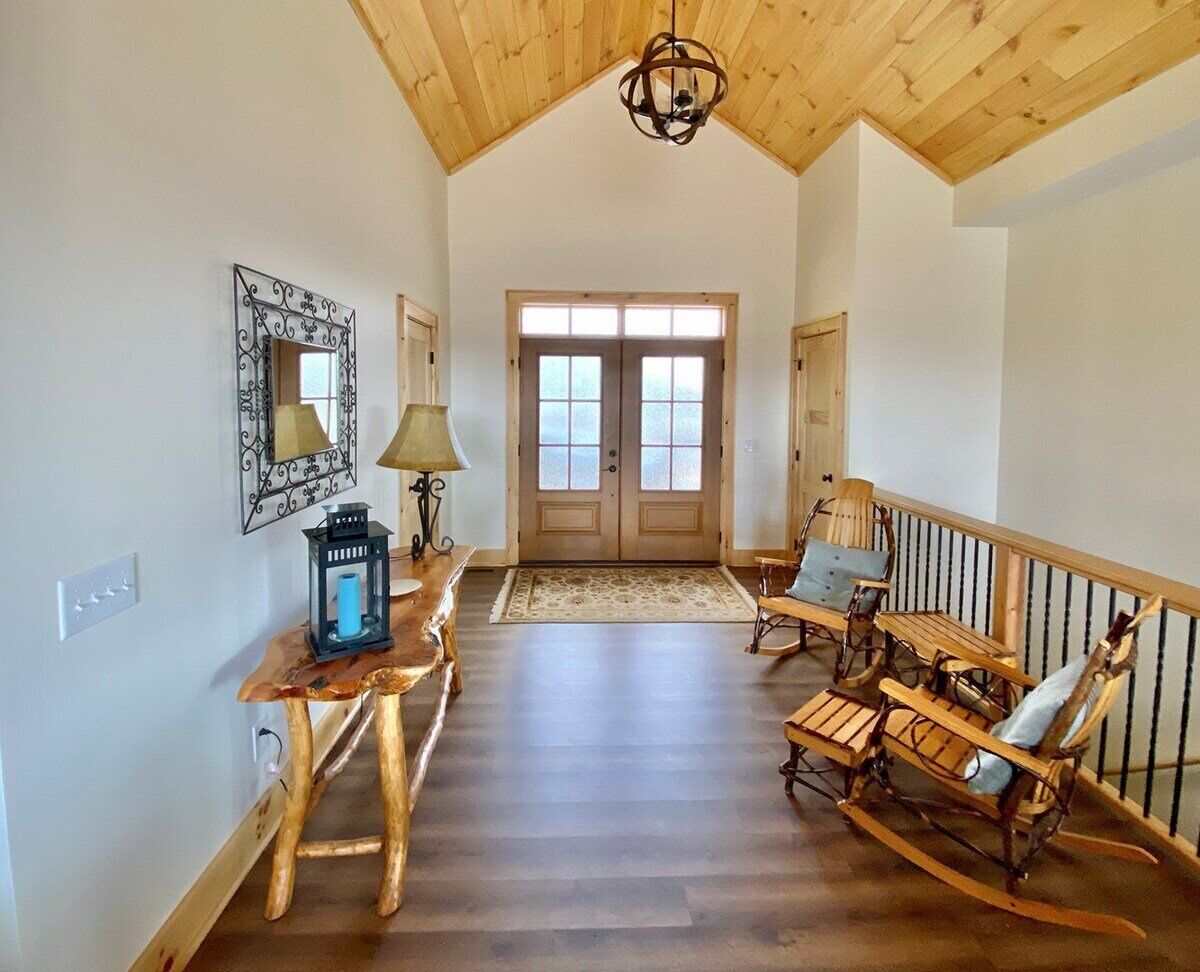 A spacious room with a high ceiling, wooden flooring, a rocking chair, a table with a lamp, and a door with windows.