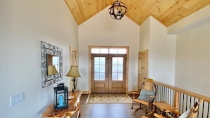 Una habitación amplia con techo alto, suelo de madera, una mecedora, una mesa con lámpara y una puerta con ventanas.