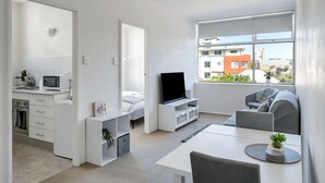A modern, well-lit apartment with a kitchenette, a dining area, a living room with a TV, and large windows offering a city view. - Quaint 1BD on the Doorstep of Manly Beach (Manly)