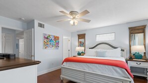 A bedroom with a bed, bedside tables, a ceiling fan, artwork on the walls, and a door leading to another room. - KeyLime Downtown Gulf View-FREE Golf Cart for 2026 (Cedar Key)