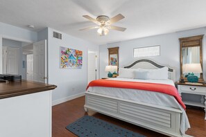 Un dormitorio con cama, mesitas de noche, ventilador de techo, obras de arte en las paredes y una puerta que da a otra habitación.