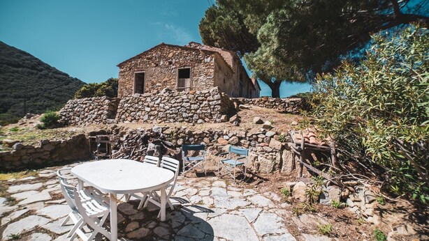 Un bâtiment en pierre doté d’un petit balcon, entouré d’arbres et agrémenté d’une terrasse en pierre avec une table et des chaises.