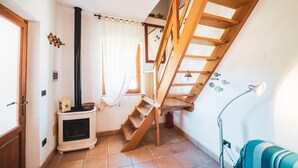 Un escalier avec des rampes en bois menant à l’étage supérieur, un poêle blanc sur lequel est posé un panier, et une fenêtre avec des rideaux blancs.