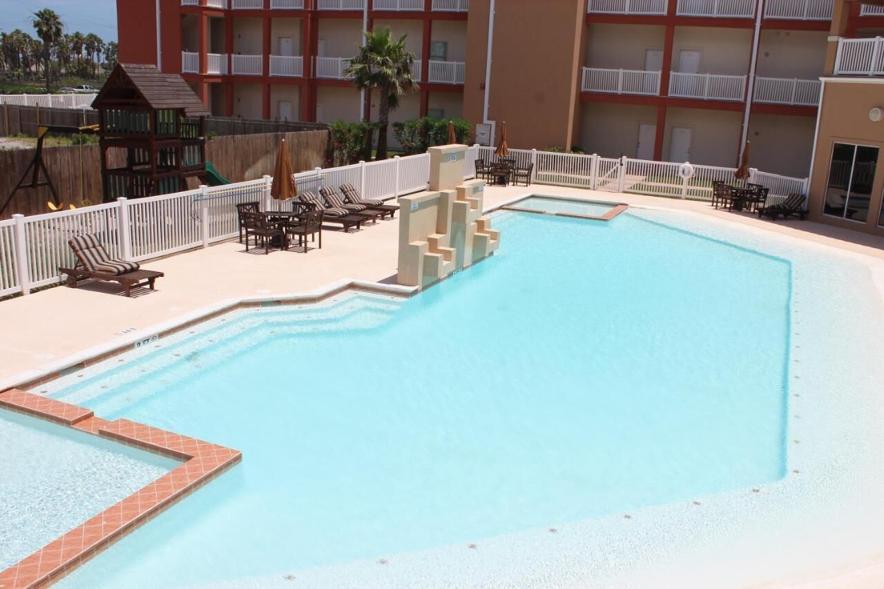 Une piscine dotée de chaises longues et d’une structure de jeux, le tout entouré d’une clôture blanche et d’un bâtiment avec des balcons.