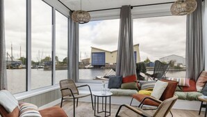 A modern living room with large windows overlooking a marina, featuring wicker chairs, a sofa with colorful cushions, and a small round table. - Harboursuite 2 incl. motorboot by Interhome (Woudsend)