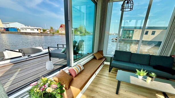 A modern room with a large window overlooking a body of water, a wooden deck, and a seating area with cushions. - Harboursuite Woudsend incl. motorboot by Interhome (Woudsend)