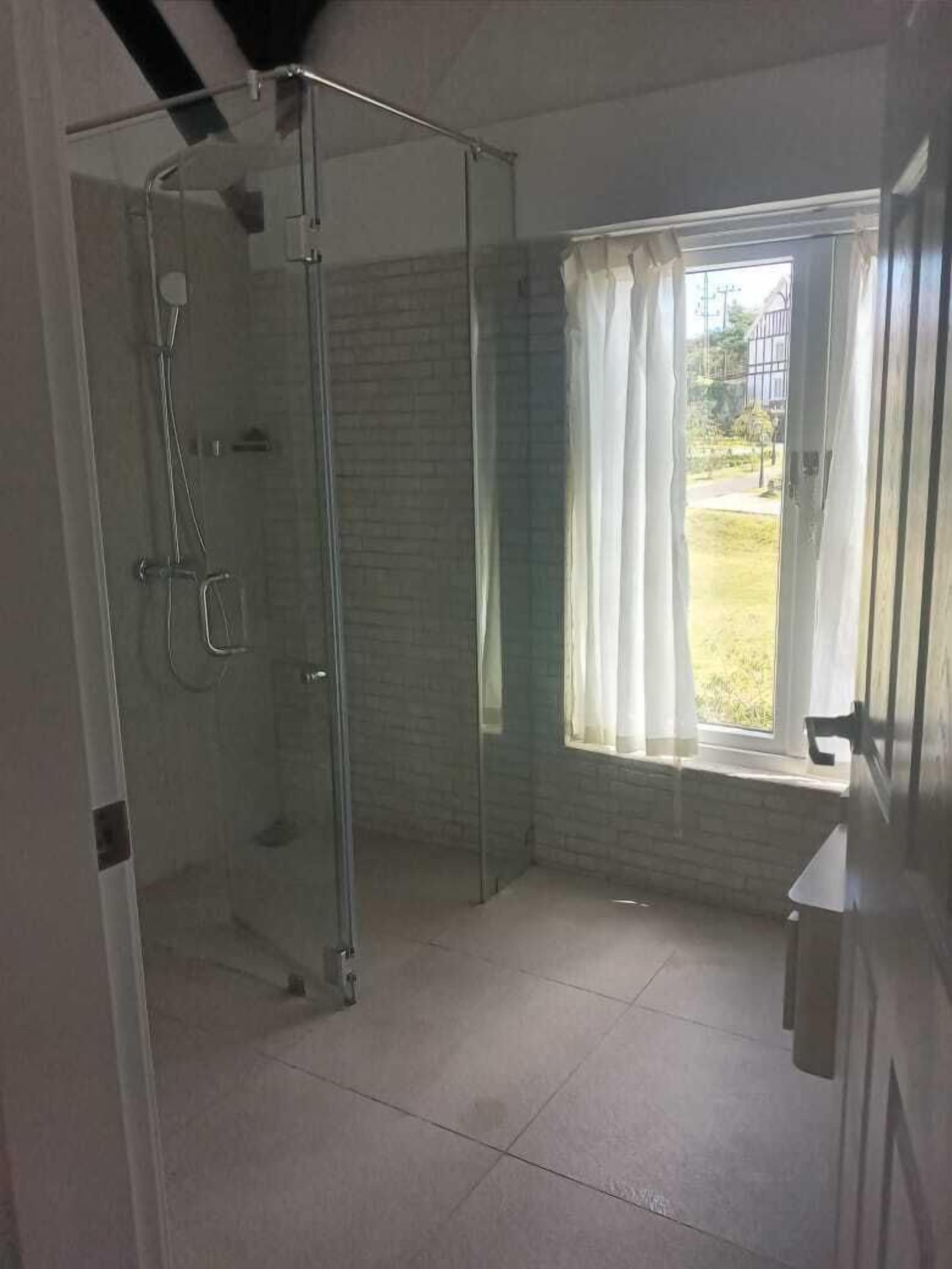 A bathroom with a glass shower enclosure, a window with curtains, and a view of the outdoors.