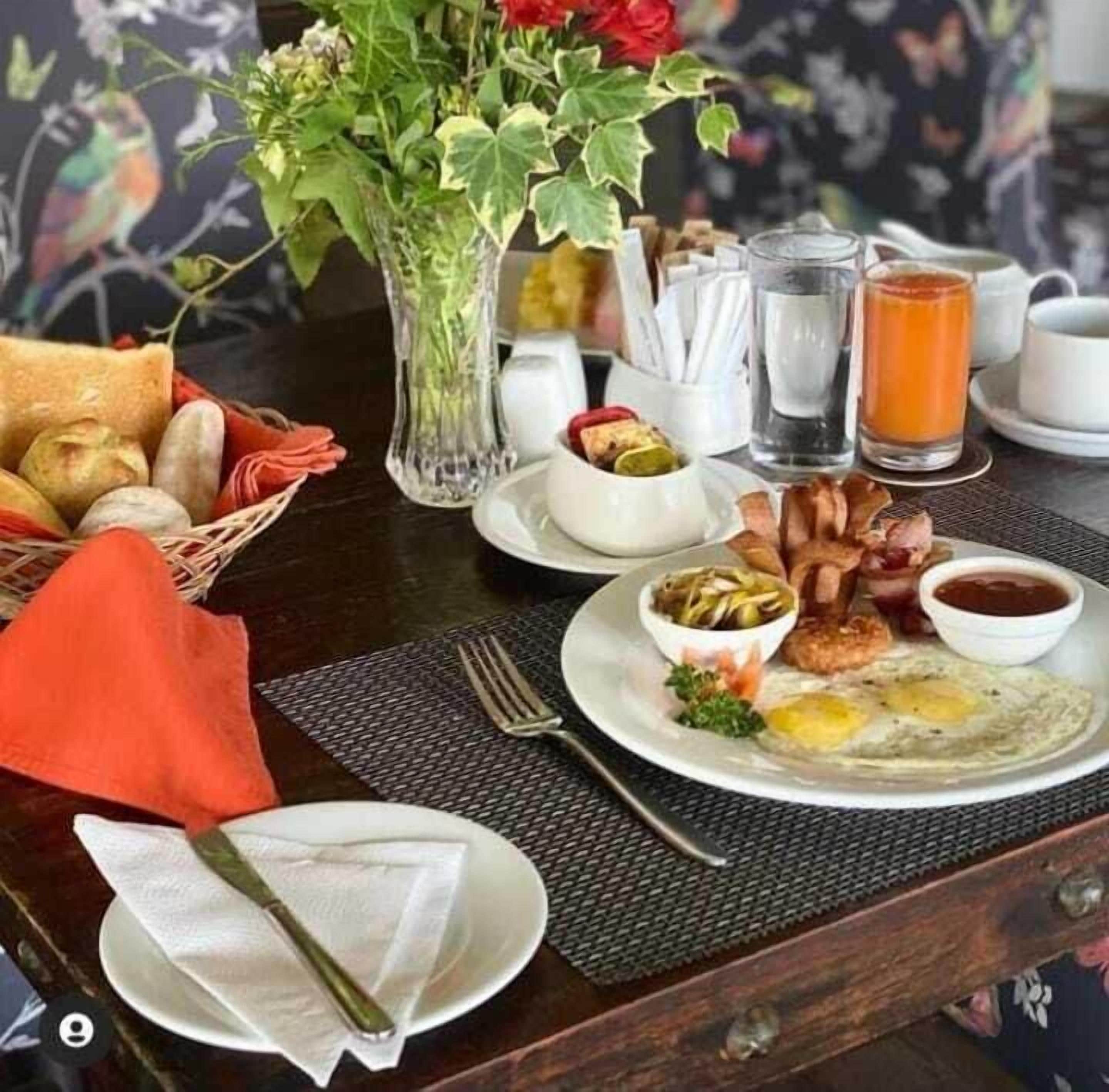 Et morgenbord med æg, bacon, en salat, en kurv med brød, en vase med blomster og et glas appelsinjuice på et bord.