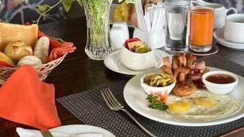 Una escena de desayuno con un plato de huevos, tocino y una ensalada, una cesta de pan, un florero y un vaso de jugo de naranja sobre una mesa.