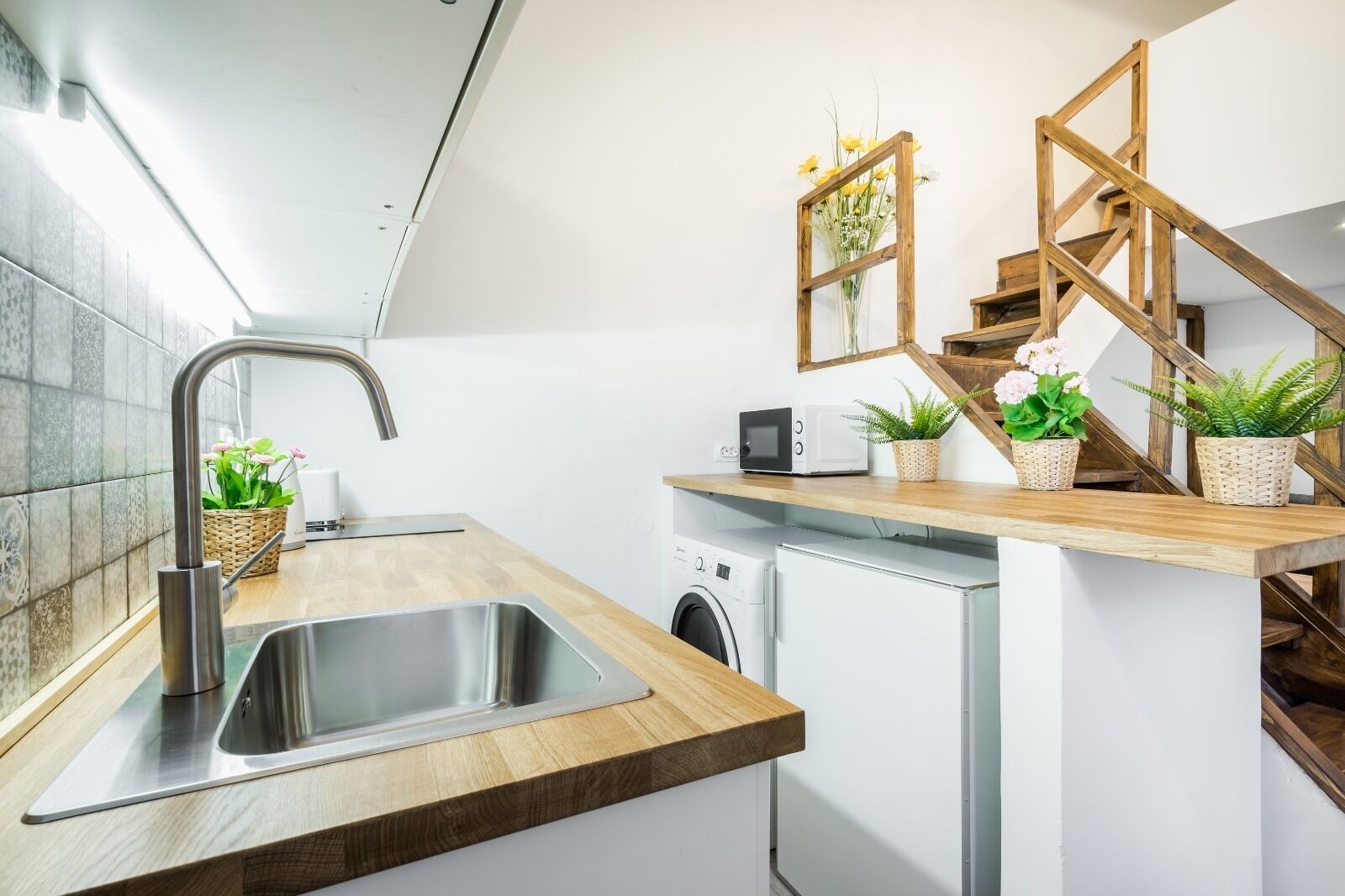 A modern kitchen with a stainless steel sink, wooden countertop, built-in washing machine, and a wooden staircase with a glass railing.