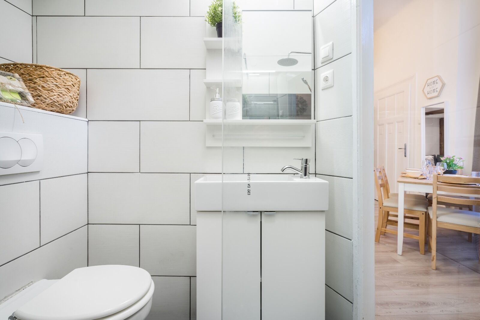 A modern bathroom with a toilet, a sink, and a shower. There is a small shelf with toiletries and a plant.