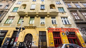 Un edificio de varios pisos con fachada amarilla, locales comerciales en la planta baja y una puerta roja.