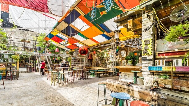 Un café al aire libre con mesas y sillas, una pared de piedra rústica y plantas colgantes.